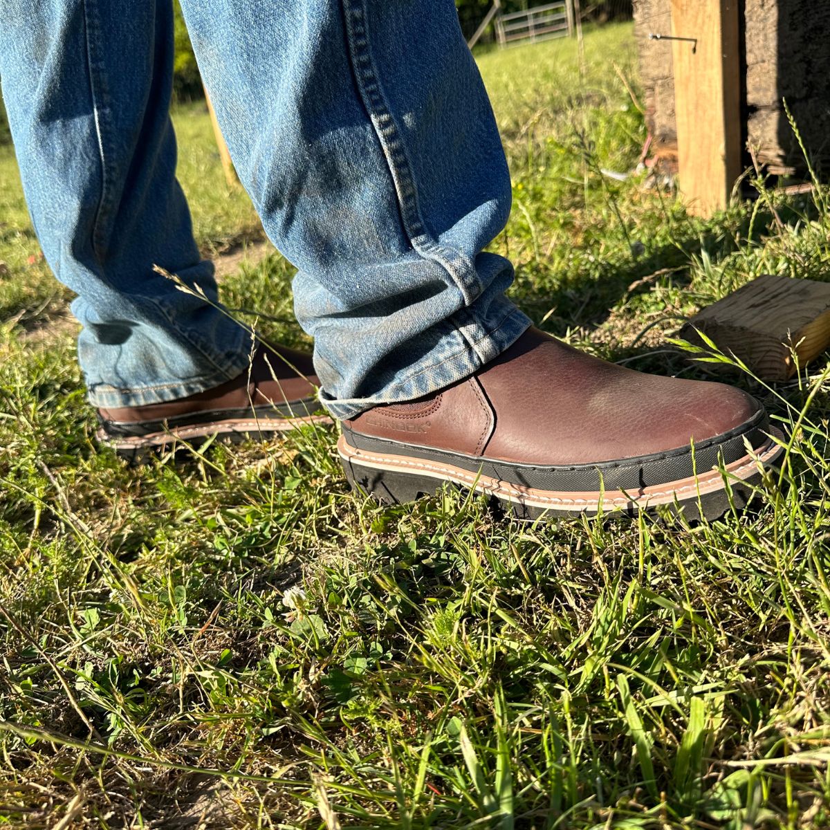 A man wearing Workhorse Romeo Men’s Light Duty Boots in Brown stands on grass. The boots feature a slip-on design with durable stitching and a rugged outsole, perfect for light-duty outdoor tasks.