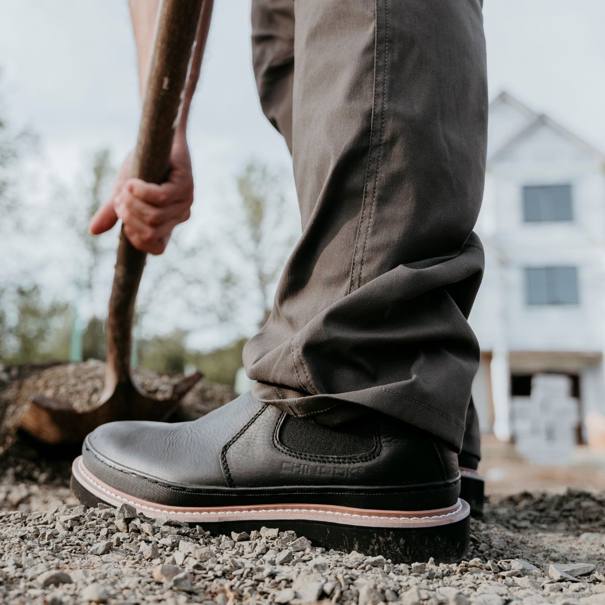 A man wearing Workhorse Romeo Men’s Light Duty Boots in Black stands on gravel while holding a shovel. The boots feature a sleek, slip-on design with durable stitching and a sturdy outsole, ideal for light-duty tasks and everyday work.