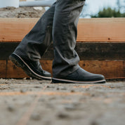 A man wearing Workhorse Romeo Men’s Light Duty Boots in Black walks on a construction site. The boots feature a slip-on design with durable stitching and a grippy outsole, ideal for light-duty work and comfortable everyday wear.