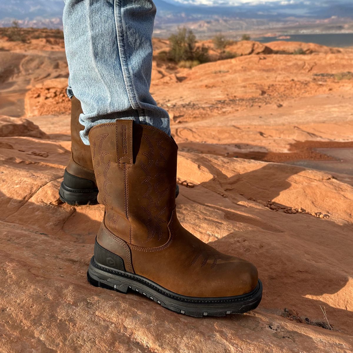 A man wearing Trailboss 11” Soft Toe Men’s Work Boots stands on rugged, red rock terrain. The boots feature durable leather with pull tabs and a sturdy outsole, built for demanding outdoor and work environments.