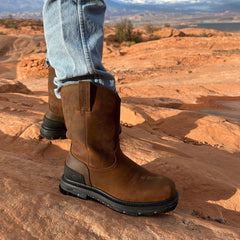 A man wearing Trailboss 11” Soft Toe Men’s Work Boots stands on rugged, red rock terrain. The boots feature durable leather with pull tabs and a sturdy outsole, built for demanding outdoor and work environments.