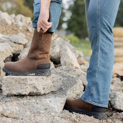 A man pulling on the Trailboss 11” Soft Toe Men’s Work Boot while standing on rocky terrain. The boot features a durable leather design with pull tabs and a rugged outsole, ideal for tough outdoor and work environments.